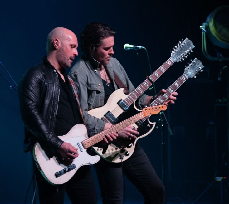 Two guitarists stand on stage side by side